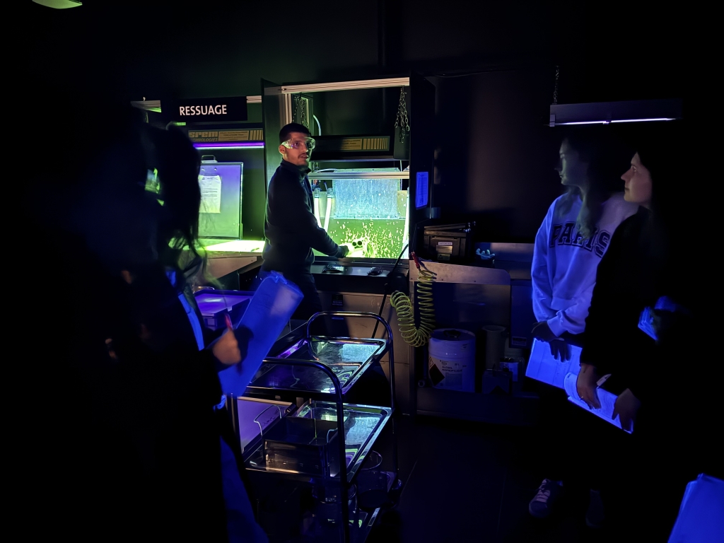 Un étudiant du Collège Galilée effectue une démonstration de technique de ressuage sous lumière UV (lumière noire) dans une cabine d'inspection. D'autres étudiants observent attentivement les résultats de ce contrôle non destructif dans le cadre du projet AIR SUPPORT.